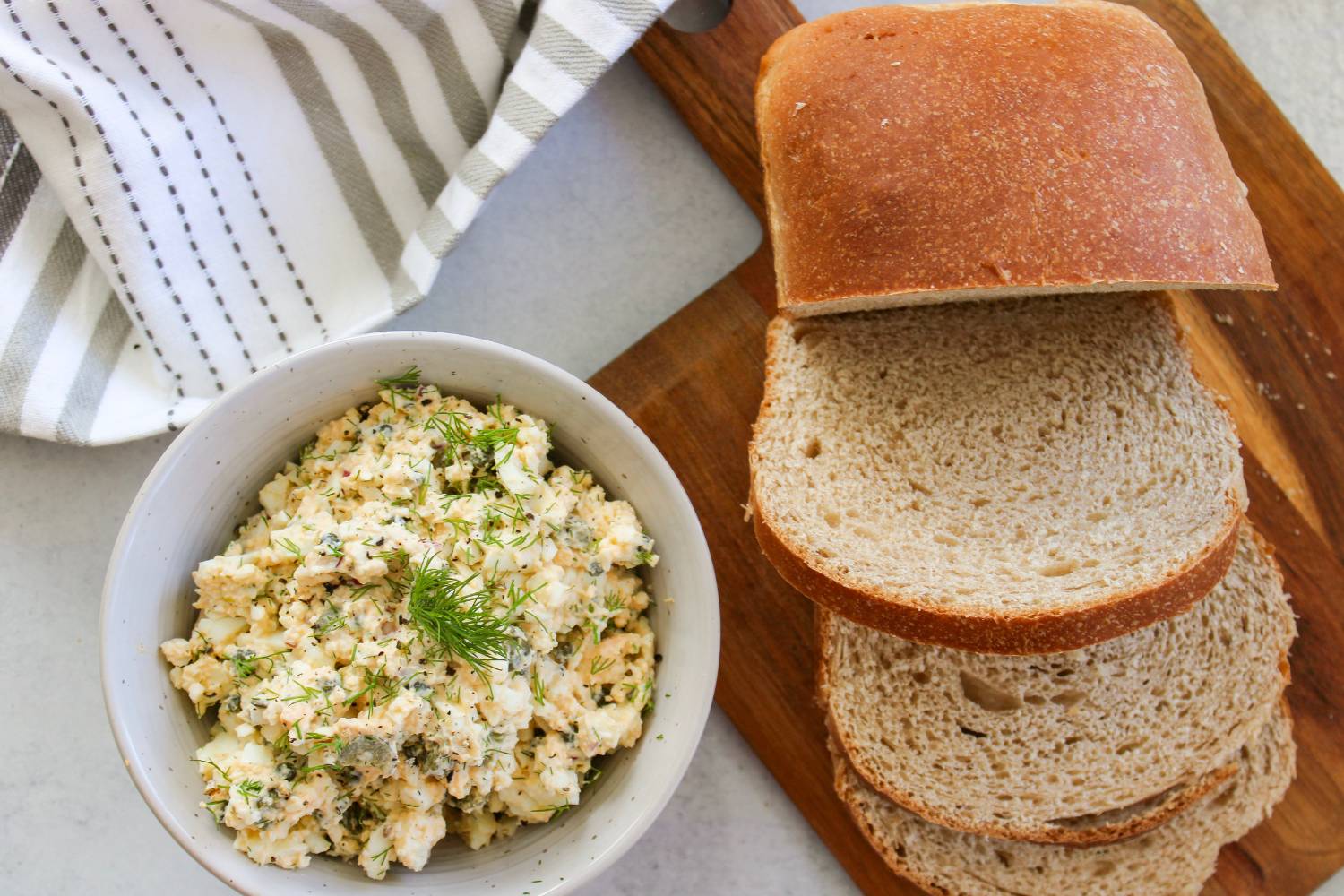 Egg salad in a bowl next to a loaf of bread on a wooden cutting board