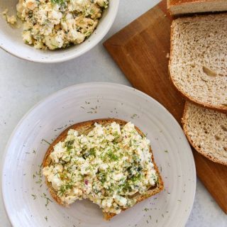 Egg salad on toast with a bite taken out next to a loaf of bread and bowl of egg salad