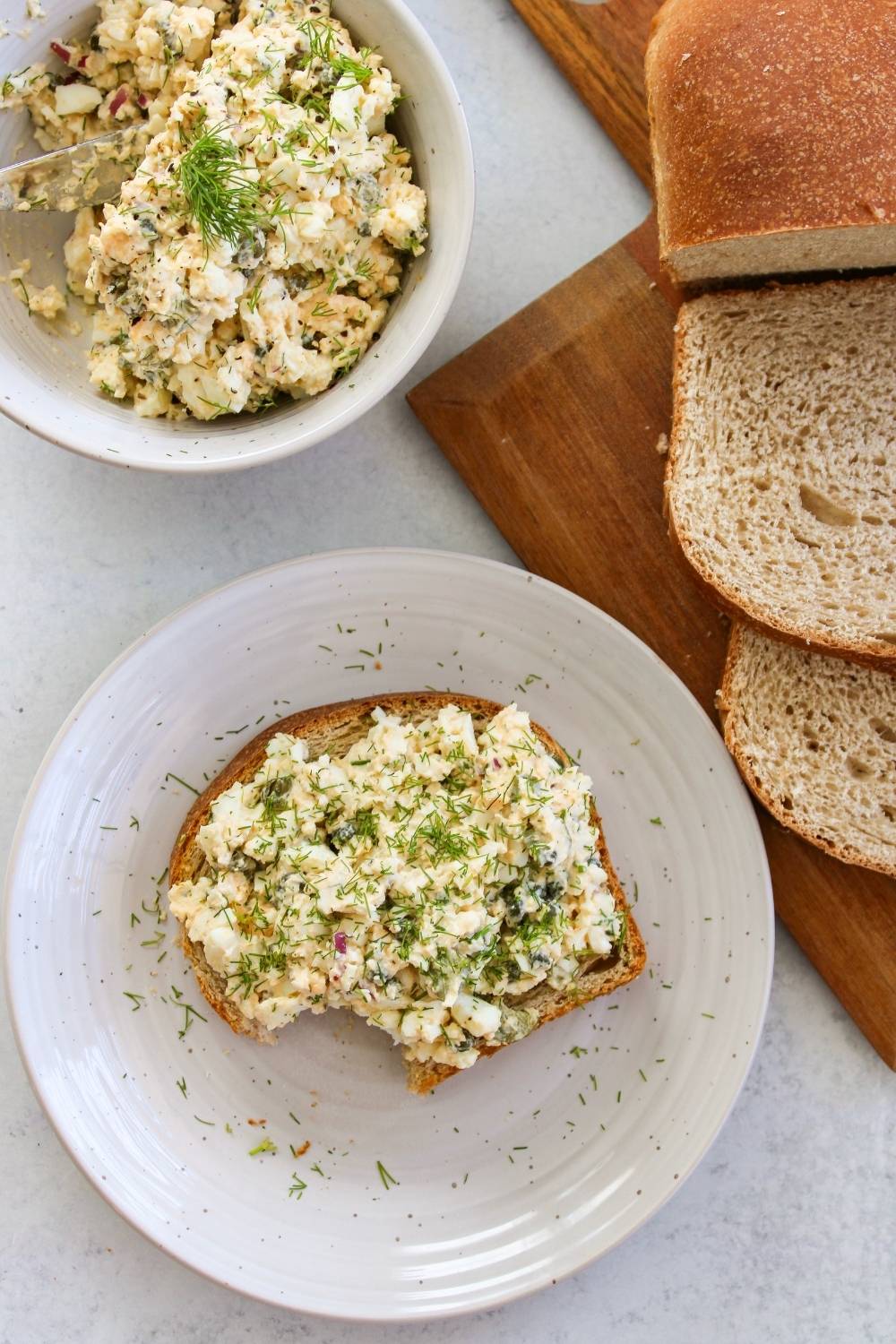 Egg salad on toast with a bite taken out next to a loaf of bread and bowl of egg salad