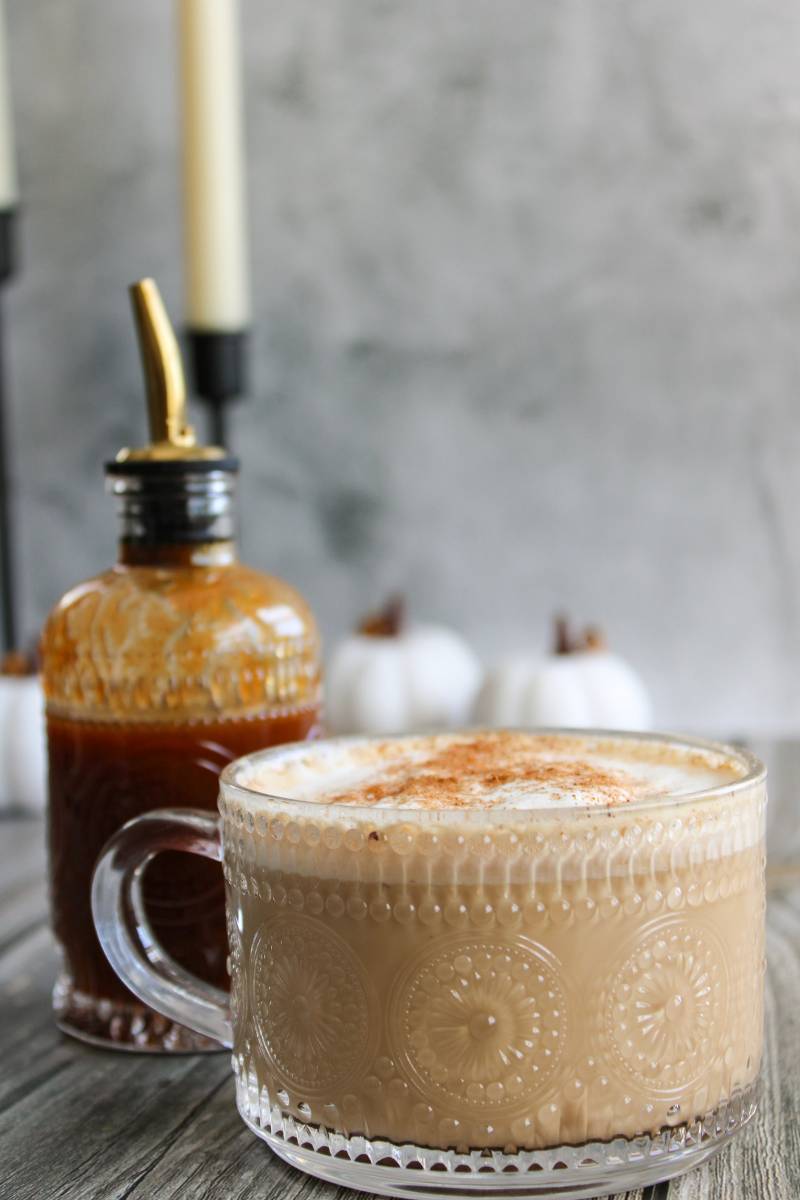 Hot pumpkin spice latte in a clear mug next to pumpkin spice syrup