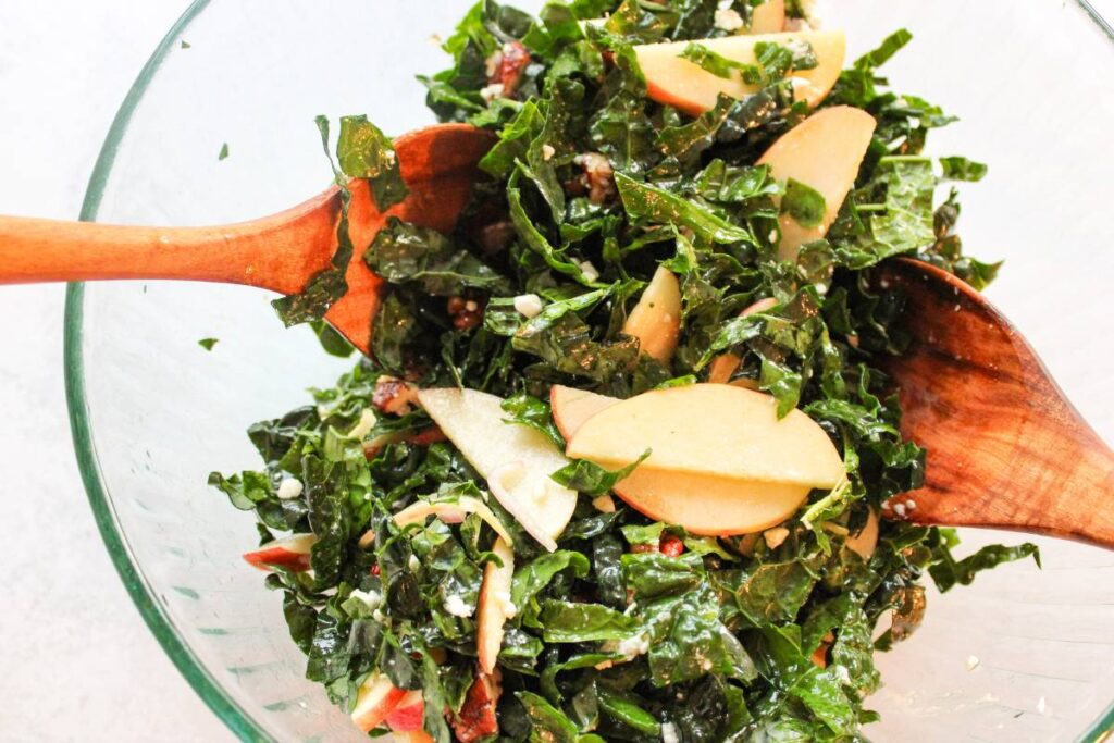 Two wooden spoons tossing kale and apple salad in a clear bowl