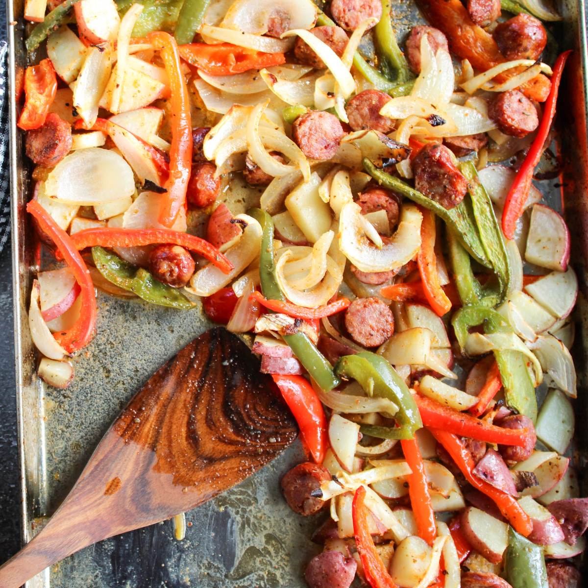 Cajun sheet pan dinner filled with Cajun Sausage, Potato and Vegetables with a wooden spatula.