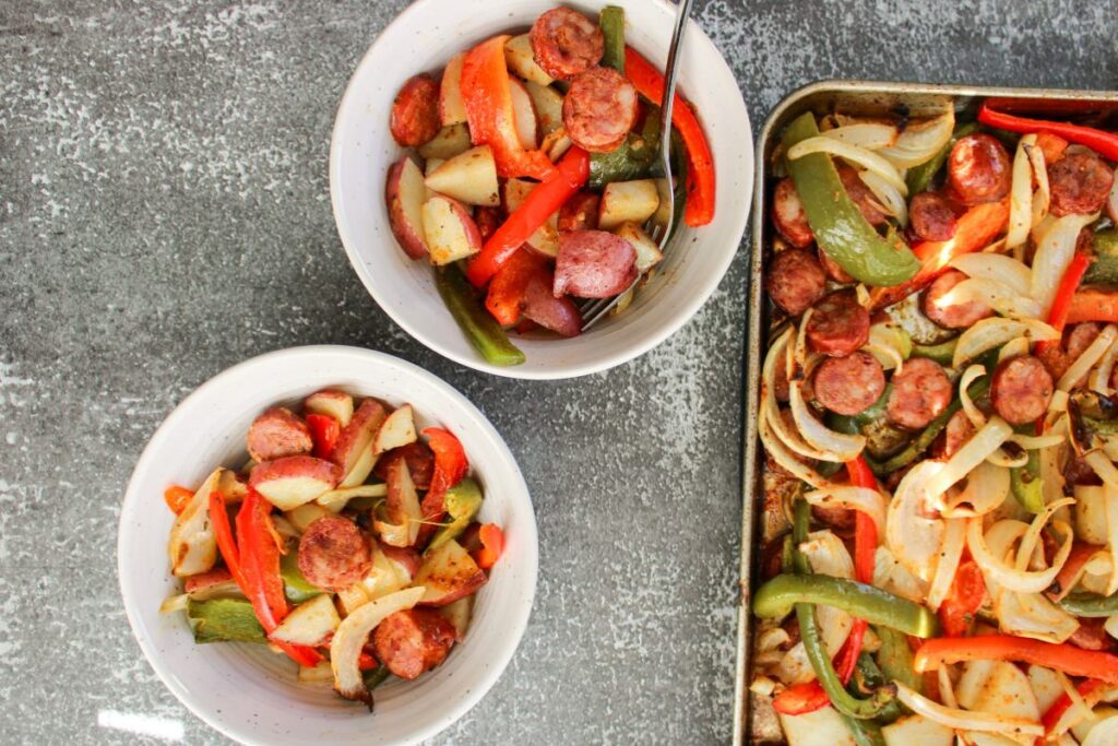 two bowls filled with Cajun Sausage, Potato and Vegetables next to a sheet pan