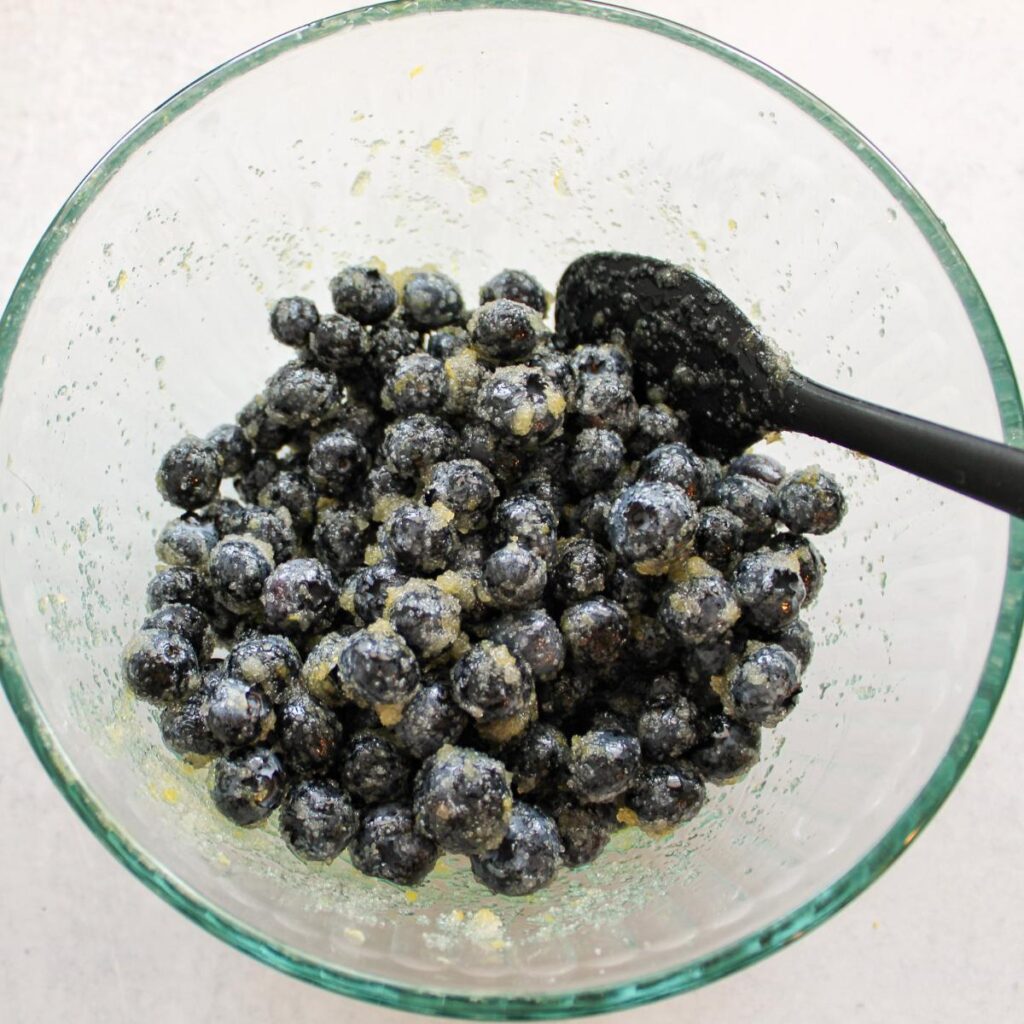 Blueberries tossed in sugar, lemon zest, and lemon juice in a clear bowl.
