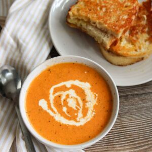 Bowl of Roasted Tomato Soup next to a plate with a grilled cheese sandwich.