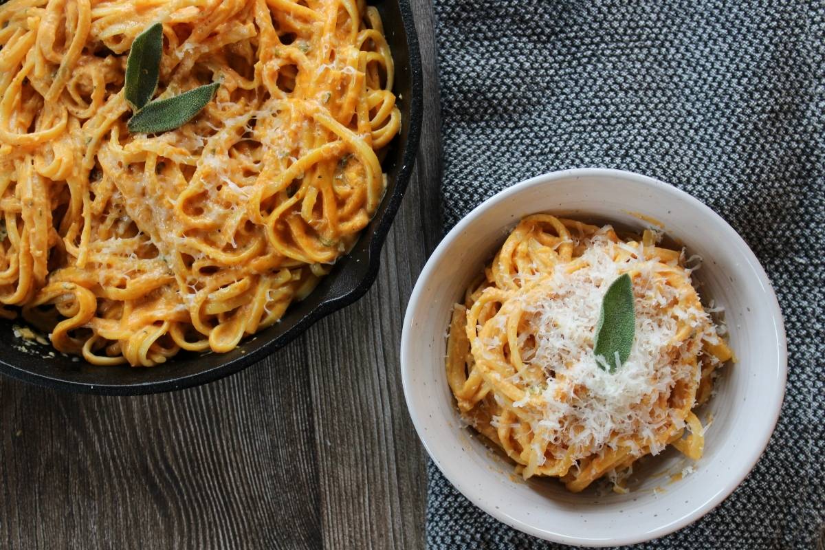 Bowl of creamy pumpkin pasta with a sage leaf on top next to a cast iron pan filled with creamy pumpkin pasta.