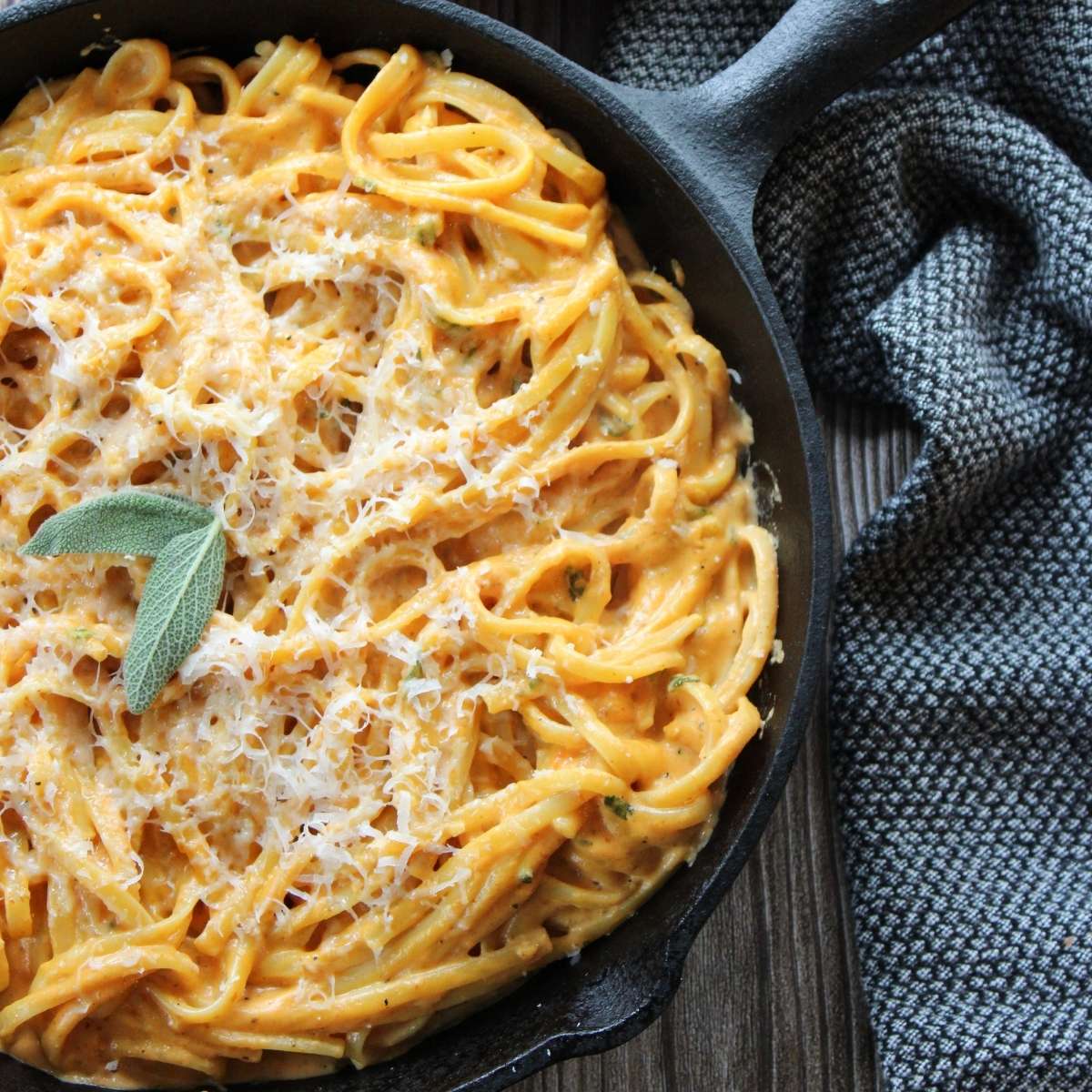 Creamy pumpkin pasta in a cast iron skillet with 2 sage leaves on top next to a black and white cloth napkin.