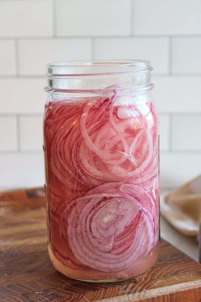 A mason jar filled with quick pickled onions on a wooden cutting board.