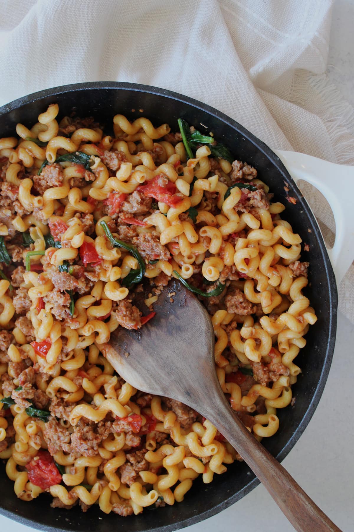 High protein pasta with Italian sausage, fresh tomatoes and spinach in a skillet with a wooden spatula.