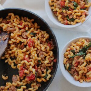 High protein pasta with Italian sausage, fresh tomatoes and spinach in a skillet next to two bowls filled with the pasta.