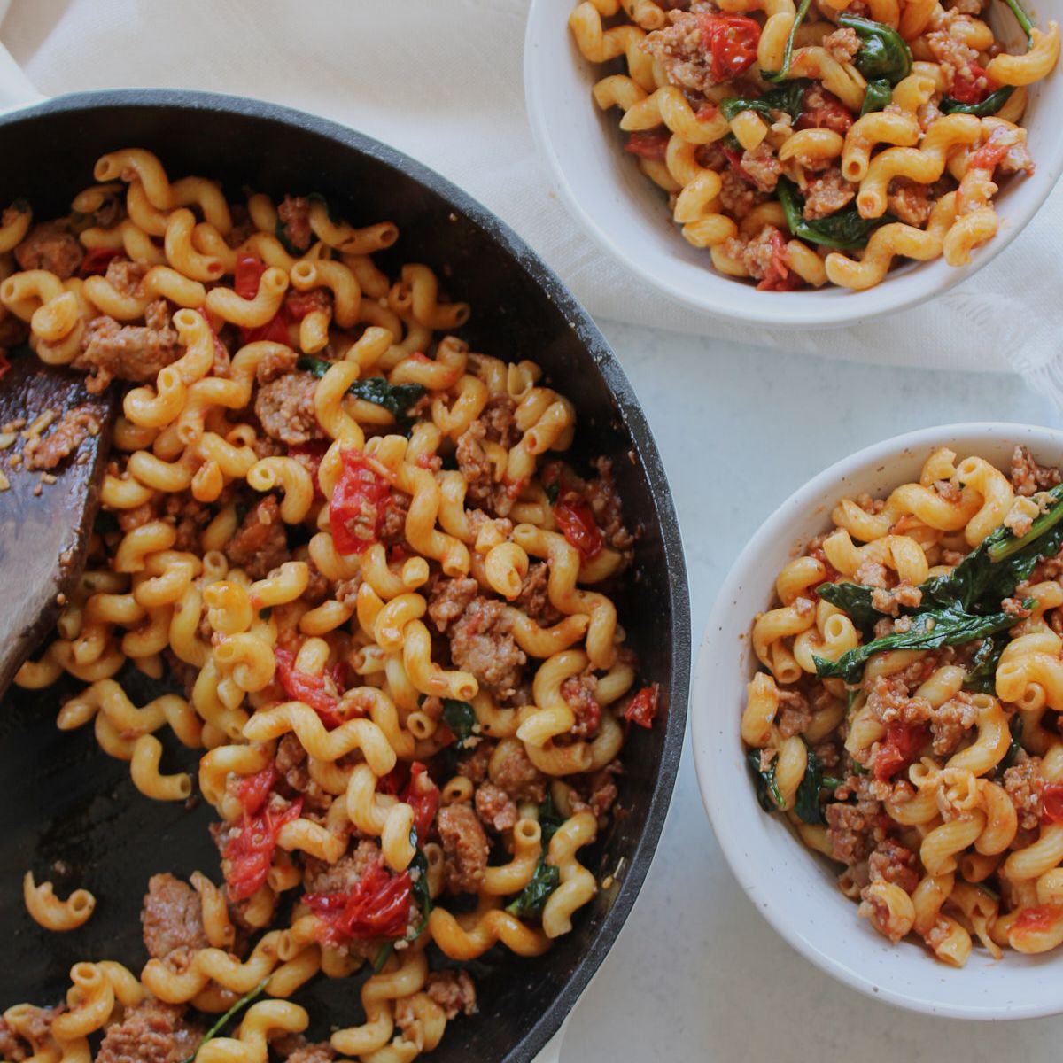 High protein pasta with Italian sausage, fresh tomatoes and spinach in a skillet next to two bowls filled with the pasta.