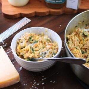 A bowl of creamy orzo next to a saucepan filled with creamy orzo next to Parmesan cheese.