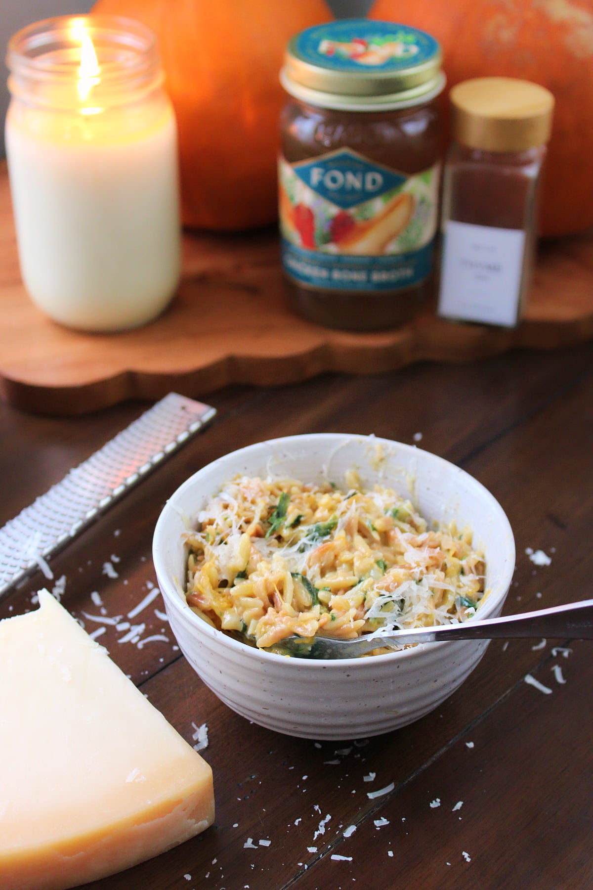 A bowl of Creamy Orzo next to Parmesan cheese and a wooden board with a candle, pumpkin, spice jar and FOND bone broth.