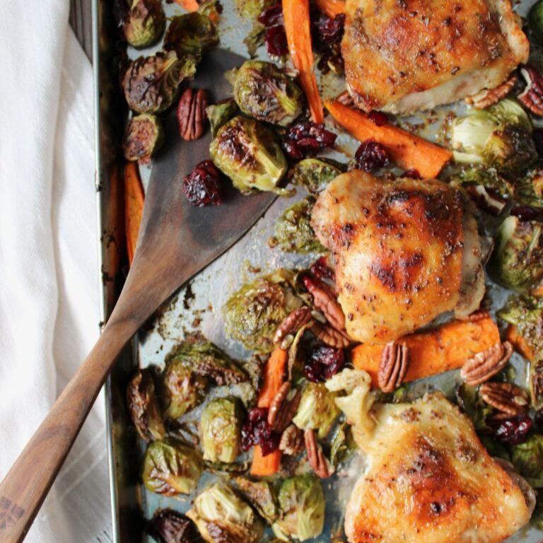 Crispy chicken thighs with caramelized Brussels sprouts and carrots tossed in a maple mustard sauce with pecans and dried cranberries on a sheet pan with a wooden spatula