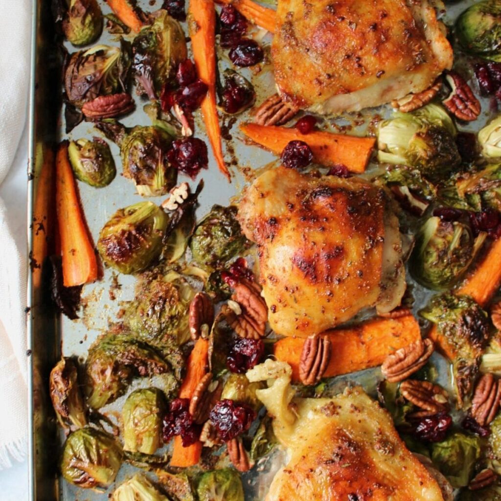 Crispy chicken thighs with caramelized Brussels sprouts and carrots tossed in a maple mustard sauce with pecans and dried cranberries on a sheet pan.